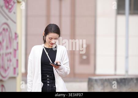 Jeune femme se tient dehors, concentrée sur son téléphone. La douce lumière du jour illumine sa douce expression. Elle porte un cardigan clair, un haut foncé et un j décontracté Banque D'Images