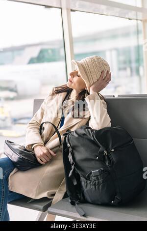 Voyageuse fatiguée se repose les yeux fermés sur un banc, sac à dos à côté d'elle dans un terminal d'aéroport occupé en attendant un départ retardé et endu Banque D'Images