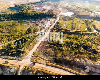 Vue aérienne d'une vaste carrière à ciel ouvert avec excavation active. Les camions traversent les routes poussiéreuses, entourés de paysages verdoyants et de bassins d'eau émergeants Banque D'Images