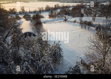 Vue aérienne d'un champ enneigé avec une clôture et un hangar au milieu des arbres d'hiver. La lumière Golden Hour réchauffe le paysage immaculé Banque D'Images