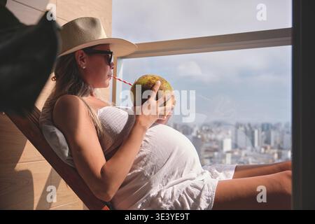 Femme enceinte appréciant des rafraîchissements et vue sur la ville sur son balcon de vacances Banque D'Images