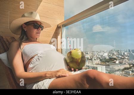Femme enceinte profitant d'une boisson tropicale et vue sur la ville depuis le balcon de l'hôtel Banque D'Images