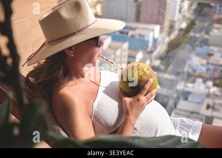Femme enceinte appréciant des rafraîchissements et vue sur la ville sur son balcon de vacances Banque D'Images