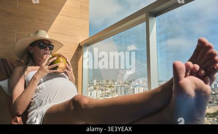 Femme enceinte profitant d'une boisson tropicale et vue sur la ville depuis un balcon ensoleillé Banque D'Images