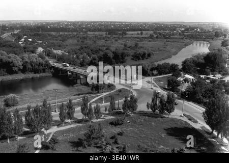 Sloviansk, milieu des années 1990, rue Svobody. Photographie de rue historique en noir et blanc prise depuis un bâtiment de 14 étages, offrant une vue dégagée en hauteur d'un pont routier central traversant la rivière. Un gros camion commercial traverse le pont vers la droite, tandis que quelques voitures de tourisme traversent l'intersection voisine entourée de grands peupliers. Un quartier tentaculaire remplit l'arrière-plan lointain. Cet authentique instantané d'archives capture parfaitement la logistique de transport post-soviétique et la vie régulière dans le Donbass paisible. Banque D'Images
