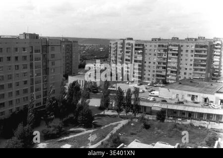 Sloviansk, milieu des années 1990, Artem Microdistrict (Lesnoy). Une photographie historique en noir et blanc capturant une vue sous un angle élevé sur une cour résidentielle caractéristique. L'espace est encadré par des blocs d'appartements à panneaux à plusieurs étages imposants des deux côtés, incarnant l'urbanisme soviétique tardif. De petits bâtiments utilitaires avec des toits plats et des véhicules domestiques stationnés occupent les zones centrales pavées au milieu des arbres verts d'été. Cet instantané d'archives réaliste dépeint avec précision la vie municipale post-soviétique et le Donbass paisible. Banque D'Images