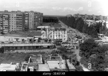 Sloviansk, milieu des années 1990, Artem Microdistrict (Lesnoy). Une photographie historique en noir et blanc montrant une vue panoramique expansive sur un secteur commercial très actif. Un marché de rue impromptu rempli de citoyens animés, de voitures et d'étals de fortune prospère le long de la large avenue. En bas au premier plan, une fondation en béton abandonnée crée un contraste frappant avec l'économie locale animée. Ce cadre documentaire détaillé préserve méticuleusement la microéconomie urbaine post-soviétique et le Donbass pacifique. Banque D'Images