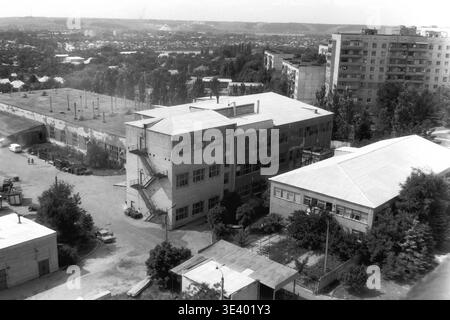 Sloviansk, milieu des années 1990, Artem Microdistrict (région de Molodiozhnaya). Une photographie historique en noir et blanc offrant une vue surélevée étendue sur une large avenue tranquille de la ville. Sur la droite, la fondation massive en béton plat d'un projet de construction inachevé se trouve sur un terrain envahi par la végétation. Une longue rangée de grands peupliers borde la route asphaltée, menant le regard vers des bâtiments municipaux lointains et un vaste paysage ouvert. Cet instantané documentaire fascinant reflète fidèlement la planification urbaine post-soviétique et la vie quotidienne dans le Donbass paisible. Banque D'Images