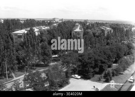 Sloviansk, milieu des années 1990, Artem Microdistrict (région de Molodiozhnaya). Une photographie historique en noir et blanc offrant un panorama panoramique panoramique d'un quartier résidentiel tranquille. Les immeubles d'appartements classiques de cinq étages sont profondément nichés dans une véritable forêt de peupliers imposants et de feuillages d'été luxuriants. En bas au premier plan, une voiture blanche suit la courbe de la route près d'un piéton marchant solitaire. Cette image documentaire hautement atmosphérique reflète parfaitement la conception civique post-soviétique et la vie régulière dans le Donbass paisible. Banque D'Images