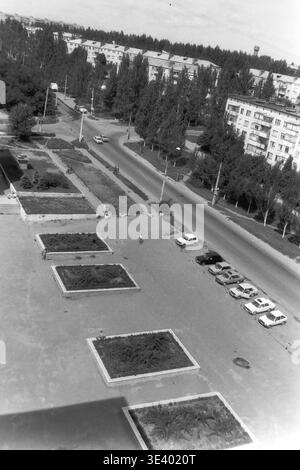 Sloviansk, milieu des années 1990, Artem Microdistrict (zone de l'Institut pédagogique). Une photographie historique en noir et blanc capturant une vaste perspective à grand angle d'une large place publique et d'une avenue adjacente. La place pavée est décorée de parterres de fleurs rectangulaires et bordée par une ligne soignée de voitures soviétiques classiques stationnées sur la droite. Un piéton seul traverse l'espace ouvert, soulignant l'atmosphère municipale calme. Cette image documentaire saisissante préserve parfaitement la conception civique post-soviétique et la vie dans le Donbass paisible. Banque D'Images