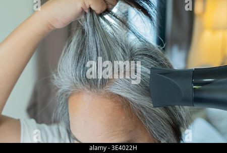 Une femme asiatique mature sèche ses cheveux gris courts avec un sèche-cheveux à la maison. Routine capillaire avec brosse et sèche-cheveux. Sèche-cheveux. Concept de soins capillaires. Coiffure w Banque D'Images