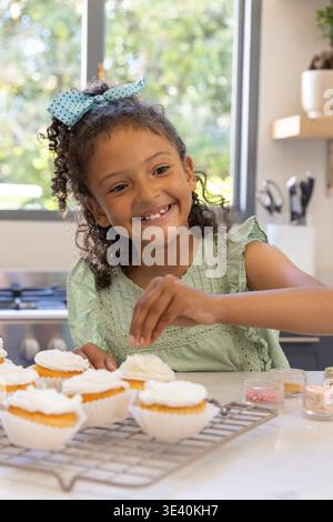 Fille enfant décorant des cupcakes sur le comptoir de la cuisine avec des saupoudres, en robe verte-dentelle et arc bleu Banque D'Images