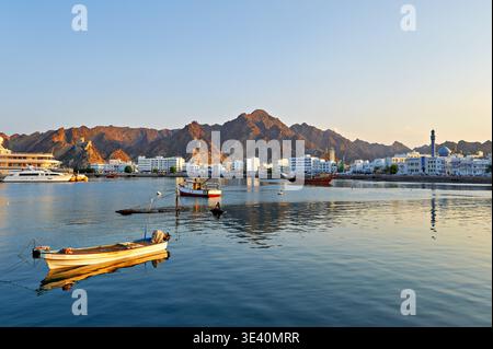 Les vagues réfléchissent la lumière du coucher de soleil à Muscat Oman tandis que les bateaux sont assis dans l'eau près du rivage. Les montagnes s'élèvent derrière les bâtiments au loin. Banque D'Images