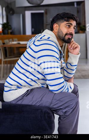 Homme indien assis sur un pouf sombre à la maison près d'une table à manger en bois, portant un sweat à capuche rayé Banque D'Images