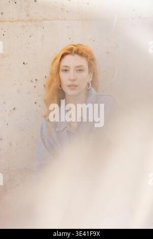 Jeune femme aux cheveux roux portant une veste en denim et des boucles d'oreilles roses debout près d'un mur Banque D'Images