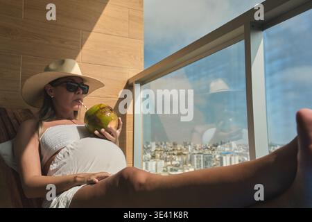 Femme enceinte se relaxant sur un balcon en profitant d'une boisson saine avec vue sur la ville Banque D'Images