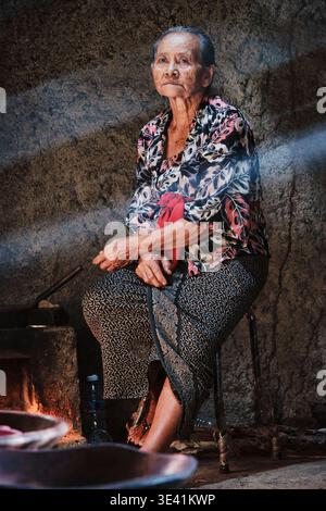 Une femme balinaise âgée est assise sur une chaise dans une cuisine simple à Bali. La lumière du matin brille à travers l'espace alors qu'elle prépare des plats traditionnels. Banque D'Images