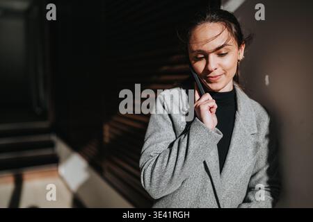 Femme d'affaires au téléphone parlant avec une équipe tout en s'appuyant contre un mur ensoleillé dans un manteau Banque D'Images
