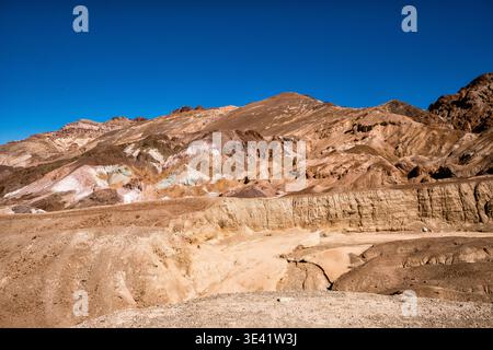 Vaste étendue désertique s'étend sous un ciel bleu clair. Les montagnes accidentées et superposées s'élèvent avec des tons terreux et rougeâtres. La lumière du soleil jaillit sur l'ancienne roc érodée Banque D'Images