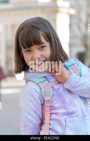Fille d'environ 8 ans avec sac à dos sourit et pose à l'extérieur. Banque D'Images
