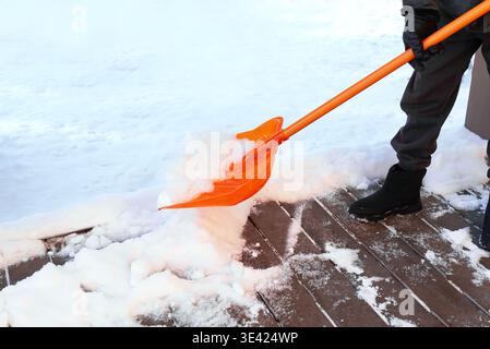 L'homme ouvrier enlève la neige de la terrasse en bois. Enlever beaucoup de neige par pelle en plastique orange en hiver froid. Banque D'Images