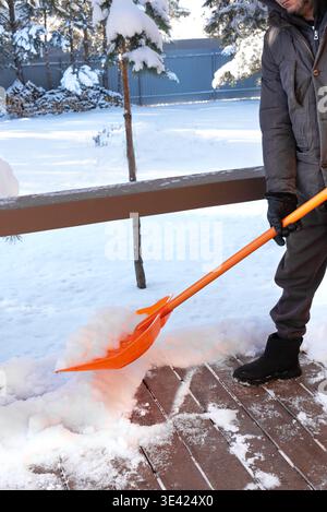 L'homme ouvrier enlève la neige de la terrasse en bois. Enlever beaucoup de neige par pelle en plastique orange en hiver froid. Banque D'Images