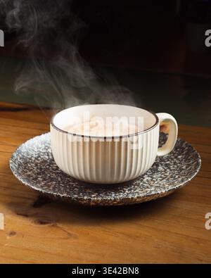 Bouclage à la vapeur à partir d'une boisson chaude dans une tasse en céramique texturée. La tasse repose sur une soucoupe mouchetée, toutes deux placées sur du bois rustique. L'éclairage doux met en valeur les subtils Banque D'Images