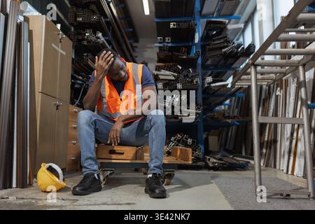 Travailleur d'entrepôt afro-américain fatigué enlevant un casque jaune et s'asseyant pour se reposer après une dure journée de travail. Banque D'Images