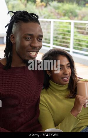 Homme afro-américain, femme indienne assise sur le pont en bois tenant une tasse tan portant des chandails en tricot Banque D'Images