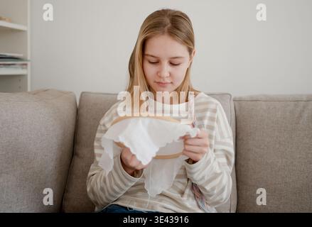 Fille assise sur Un canapé à la maison apprenant à broder sur Un cerceau Banque D'Images
