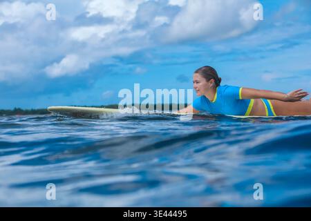 Jeune femme pagayant sur l'enseignement de planche de surf, profil latéral avec geste de guidage et expression calme, petite houle Banque D'Images