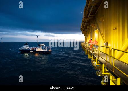 Travailleurs offshore sur une passerelle de plate-forme éclairée surplombant un navire de soutien dans un parc éolien au crépuscule, avec des turbines à l'horizon des nuages sombres de tempête Banque D'Images