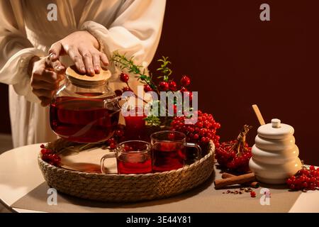 Jeune femme versant du thé viburnum chaud de la théière en verre dans la tasse à la maison Banque D'Images
