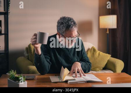 Un homme mûr avec des lunettes est assis à un bureau en bois dans un salon chaleureux, examinant son emploi du temps dans un planificateur tout en tenant une tasse de café, illustrant Banque D'Images