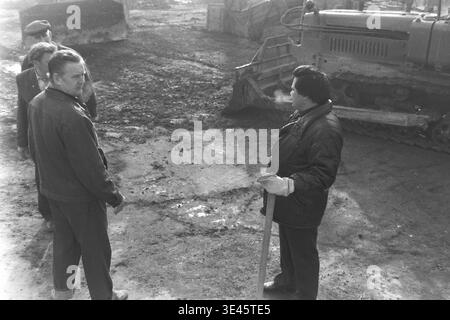 Sloviansk, avril 1989, cour d'usine AIZ. Deux directeurs d'usine discutent activement du processus de travail en cours à côté d'un bulldozer à chenilles industriel lourd lors d'une journée de nettoyage de bénévoles de printemps. Vêtus de vêtements d'extérieur chauds, les hommes planifient soigneusement les opérations de défrichement du territoire dans la cour de fabrication remplie de saleté et de débris. Cette scène extérieure franche capture merveilleusement le leadership dévoué, le travail d'équipe et les fortes valeurs de service communautaire dans le magnifique Donbass paisible. Banque D'Images