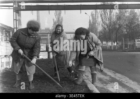 Sloviansk, avril 1989, cour d'usine AIZ. Un groupe de trois femmes dévouées portant des manteaux chauds et des foulards effectuent des travaux manuels lors d'un événement traditionnel de nettoyage de bénévoles subbotnik au printemps. Ils utilisent activement des pelles pour creuser et préparer le sol le long d'une bordure de route sur le territoire industriel, probablement pour la plantation. Cette scène très authentique met en évidence la contribution essentielle des travailleuses à l’entretien et à l’embellissement du territoire de l’entreprise dans le Donbass paisible. Banque D'Images