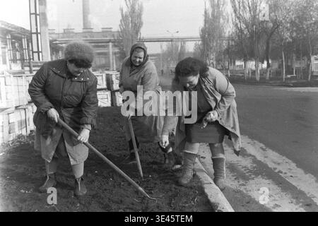 Sloviansk, avril 1989, cour d'usine AIZ. Un groupe de trois femmes dévouées portant des manteaux chauds et des foulards effectuent des travaux manuels lors d'un événement traditionnel de nettoyage de bénévoles subbotnik au printemps. Ils utilisent activement des pelles pour creuser et préparer le sol le long d'une bordure de route sur le territoire industriel, probablement pour la plantation. Cette scène très authentique met en évidence la contribution essentielle des travailleuses à l’entretien et à l’embellissement du territoire de l’entreprise dans le Donbass paisible. Banque D'Images