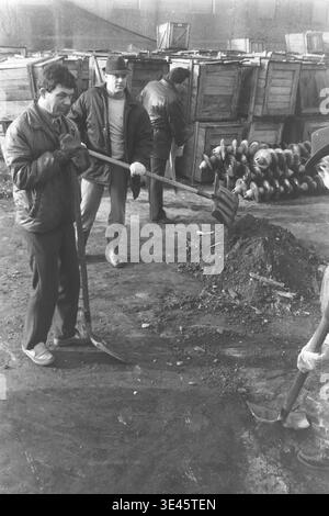 Sloviansk, avril 1989, cour d'usine AIZ. Trois hommes de l'équipe de direction de l'usine collaborent activement pour déplacer un lourd tas de saleté et de débris à l'aide de longues pelles lors d'une journée de nettoyage de bénévoles de printemps. Ils travaillent dur dans la cour industrielle entourée de caisses en bois et de grands isolants électriques en céramique. Cette photographie extérieure engageante illustre magnifiquement le travail d'équipe physique unifié et les fortes responsabilités de la communauté dans le Donbass paisible. Banque D'Images