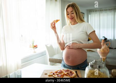 Femme enceinte heureuse mangeant de la pizza à la maison. Envies de grossesse, mode de vie de maternité, nutrition et concept de bien-être prénatal. Banque D'Images