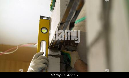 Gros plan d'un travailleur de la construction en gants tenant un niveau à bulle jaune contre un mur de blocs de béton. Vérification de l'alignement vertical précis et d'un Banque D'Images