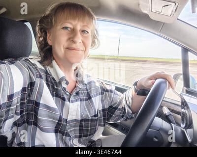 Femme prend une pause dans sa voiture, souriant à la caméra, portant une chemise à carreaux, profitant de son voyage par une journée ensoleillée. Banque D'Images