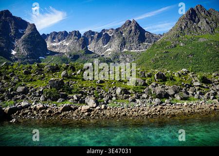 Chaîne de montagnes rocheuses s'élevant au-dessus de la vallée verte et du rivage d'eau claire sous le ciel bleu avec des nuages dispersés dans un paysage éloigné. Banque D'Images