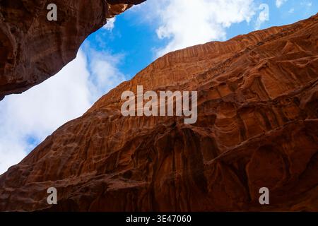 Red sandstone canyon walls rise steeply toward blue sky with clouds, viewed from below inside a narrow rocky desert gorge. Banque D'Images