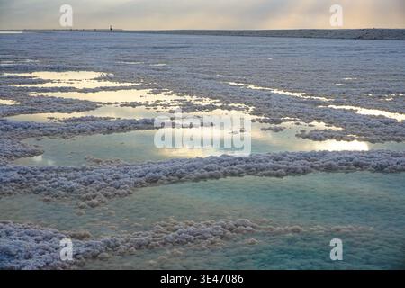 Formations salines et bassins d'eau peu profonds sur le rivage de la mer morte reflétant le ciel nuageux au coucher du soleil, créant un paysage minéral texturé en Jordanie. Banque D'Images