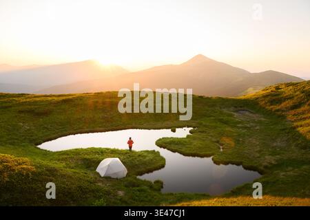 Petit lac alpin reflétant la lumière du lever du soleil entouré d'herbe verte et de sommets lointains. Paysage paisible de haute altitude avec des tons doux et chauds et une sérénité naturelle Banque D'Images