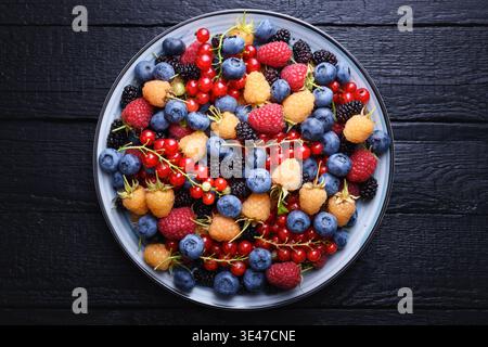 Vue de dessus de baies assorties dans un bol bleu sur fond de bois foncé. Mélange de fruits de saison, récolte biologique sans cerises. Pose à plat Banque D'Images