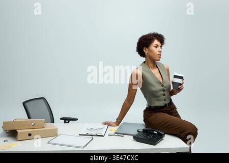 Charmante jeune femme dans une tenue élégante s'assoit en toute confiance à un bureau, sirotant un café réfléchi. Banque D'Images