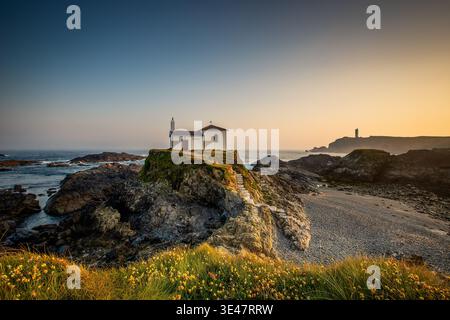 Chapelle solitaire sur la côte rocheuse surplombant l'océan au coucher du soleil avec phare lointain et chemin, Valdovino, Espagne Banque D'Images