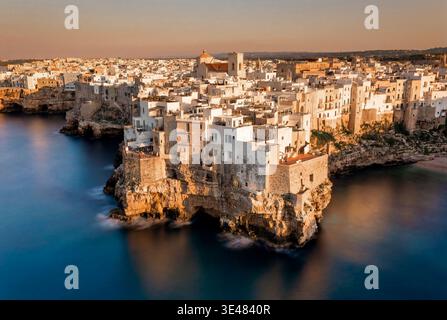 Vue aérienne des bâtiments blancs historiques perchés sur des falaises de calcaire surplombant la mer turquoise pendant le coucher du soleil à Polignano a Mare, Pouilles, Italie Banque D'Images