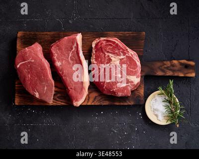 Trois morceaux de bœuf cru sont disposés sur une planche à découper en bois. Ils comprennent le Rib-eye, le rôti de bœuf et le flanc. Un petit bol de sel et d'herbes fraîches est Banque D'Images